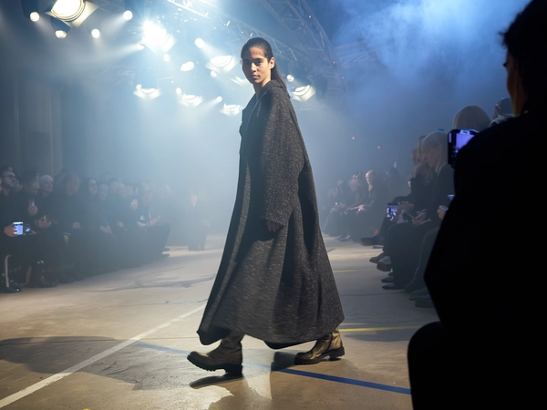 A lone female model drapes an oversized, textured coat that sways voluminously around her mid-stride, her heavy boots pressing into a fog-softened runway. Beams of sharp, white stage lights slice through the mist, casting long, diffused rays that silhouette her form with a ghostly glow. The catwalk, bordered by darkened guests blurring into shadow, reveals subtle concrete floor scuffs and painted line markers. Ambient haze mingles with sensor grain and a soft bloom around overhead tungsten fixtures, while the coat's fabric weave and the matte sheen on weathered leather boots pop vividly. Shot from a low diagonal by the aisle, the camera captures a slightly skewed frame where the fog curls upwards, the model’s focused expression piercing through atmospheric softness—contemporary runway snapshot, shot on iPhone