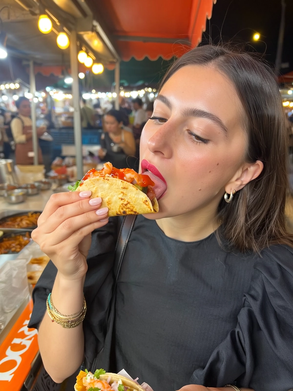 Under the cool hum of lanterns at an outdoor night market, a brunette beauty with sleek hair tucked behind one ear enjoys a moment of poised indulgence. Her matte lipstick, a vibrant shade echoing the fiery red salsa, contrasts elegantly against her smooth skin and understated makeup. She wears a soft, black cotton blouse with subtle puff sleeves, lending contemporary-chic balance to the lively backdrop.

In mid-bite, she lifts a shrimp taco delicately toward her parted lips, the crisped edges of the toasted tortilla cupped perfectly. The bright salsa glistens with a glossy sheen, its deep ruby droplets catching the subtle glow from strings of warm-hued bulbs. Her eyes are half-lidded, emanating a serene calm, while the precise angle of her bite highlights the succulent shrimp and fresh cilantro threads nestled inside.

The night air carries a faint smoky warmth from nearby grills as the blurred market stalls flicker in the background, their colors softened by shallow depth. The texture of her shirt’s fine weave is palpable, echoed by the tactile contrast of her glossy lips and the slight crunch of taco edges. The composition, a casual iPhone shot taken at arm’s length with a casual tilt, captures the moment’s vibrant authenticity with micro-contrasts and gentle clipping that enrich the warm palette.

This snapshot fuses effortless high-fashion allure with the energetic spirit of street food culture, each element layered with textural detail yet held in a composed tranquil expression —perfectly resembling a candid iPhone photo.
