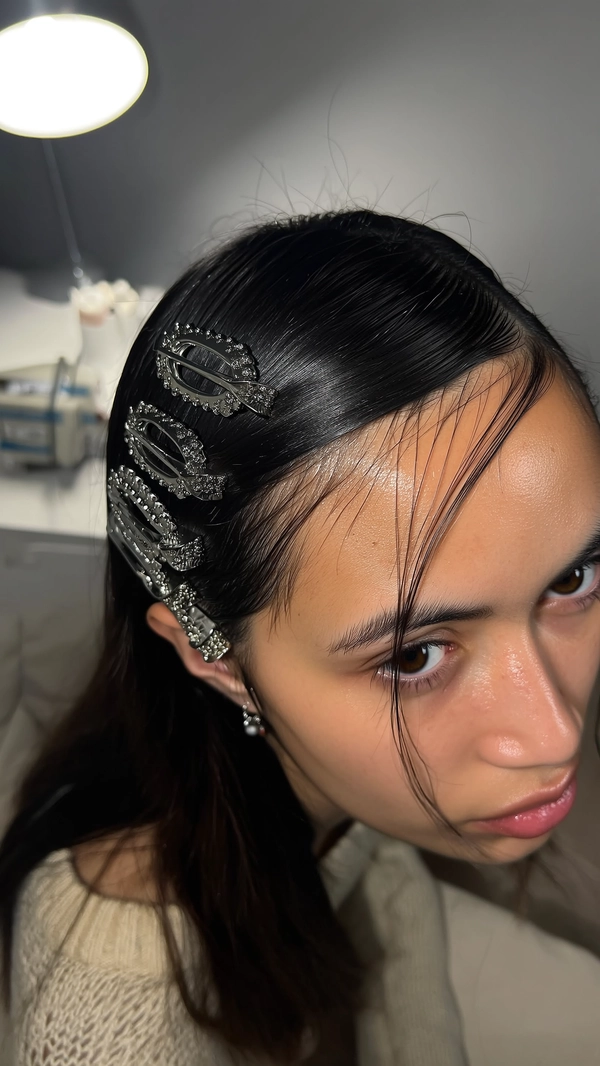 Matte metal barrettes stack like a sleek ladder along a deep side part, their textured surfaces catching a muted glow from a softly diffused bedroom desk lamp. A calm-faced girl meets the gaze directly, her glass-skin glow subtly shimmering, while fly-away strands trace gentle shadows across her forehead. The close frame skews slightly high-angle, capturing delicate pores and the fine fuzz of her knit collar peeking into the shot. Background hints of city pavement blocks blur faintly, grounding this intimate moment of stillness and style perfectly—authentic iPhone close-up, statement hair-clip focus
