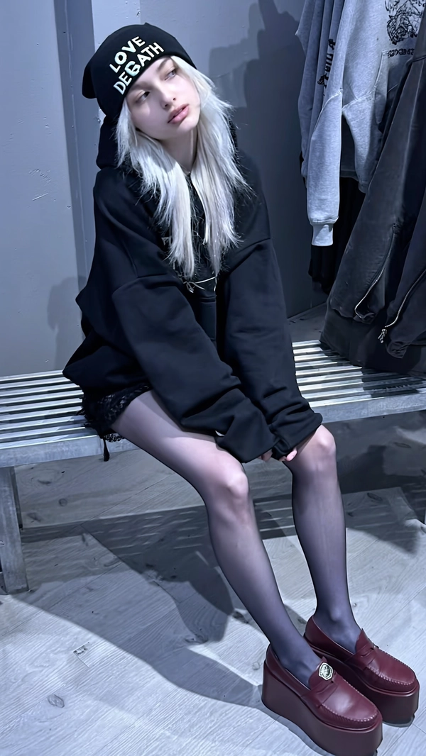 A candid mid-2010s-style snapshot featuring a pale young woman with icy platinum hair styled casually loose, seated on a metal bench inside a monochrome concept store. She wears a huge black hoodie, sheer tights, and maroon platform creepers, complemented by a beanie embroidered with “LOVE AND DEATH.” The subject’s relaxed expression gazes off to the side, conveying subtle, ambiguous emotion. The lighting is cold and matte with soft shadows stretching along a wooden floor, intentionally exhibiting muted color saturation, softened contrast, and distinctly cool-toned bluish-gray shadows. Visible textures include realistic skin details, detailed fabric grain of the hoodie and tights, individual icy hair strands, and clear accessory textures. The framing is slightly off-center and casually tilted, capturing spontaneous intimacy and informal snapshot aesthetics characteristic of mid-2010s casual youth photography.
