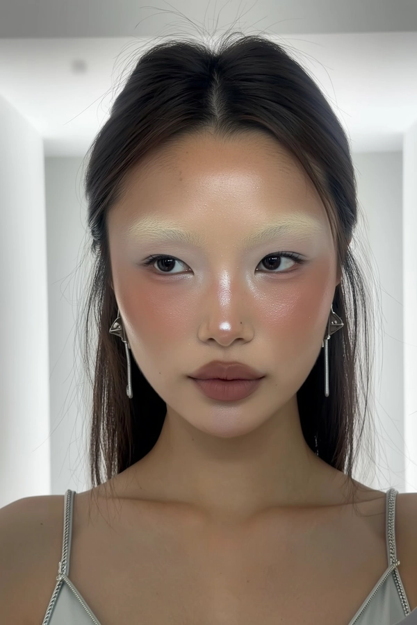 A close-up, authentic iPhone selfie-style portrait of a young woman with naturally bleached eyebrows softly blending into her skin, showcasing sculpted cheeks with subtle shimmering highlight that adds dimension. Her makeup remains natural with muted matte lips and minimal eye makeup emphasizing faint icy or silver tones. The lighting is sterile and soft, casting gentle, diffused shadows that highlight the shimmer on her skin while keeping the overall mood calm. She gazes neutrally but engagingly into the camera, her hair naturally framing her face. The background is a smooth, seamless white environment without visible corners, evoking a minimalist, modern aesthetic akin to a softly lit white cube. She wears a simple, light-colored top with understated silver accessories, reinforcing the effortless and contemporary vibe of the iPhone selfie.