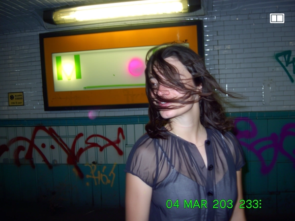 In the harsh pop of the built-in flash, a woman stands at the subway entrance, her sheer blouse catching quick folds as the evening wind flutters loose strands of hair around her face. The pixelated glow from the metro sign blurs softly behind, tinted by mixed fluorescent and tungsten lights, casting pastel washes on graffiti-smeared tiling. The camera jitters in a slight Dutch tilt, cropping the frame oddly, while speckles of CCD noise and soft edges smear faintly. The timestamp in blocky green reads “04 MAR 2003  23:18,” a subtle battery icon bounces in the corner. The photo quality is low-resolution and somewhat grainy, freezing this fleeting urban moment with casual charm. —early-2000s digicam snapshot, grainy low-res quality
