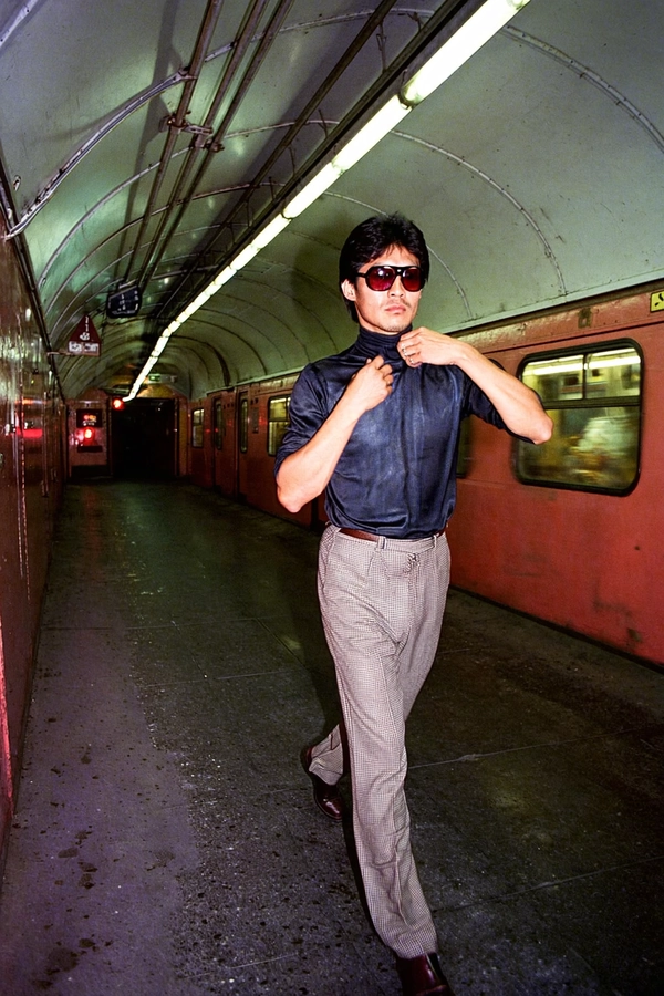 In a gritty, shadow-dappled subway tunnel from the late 1970s, a Southeast Asian man strides with deliberate confidence, his flared checkered trousers catching the flickering neon light. The tunnel’s grimy tiled walls rebound cool blues and electric greens off the glossy lenses of his oversized, tinted sunglasses, each reflection hinting at blurred graffiti and steaming vent pipes. His thick black hair is perfectly coiffed in a neat wave, a slight sheen betraying the effort in styling, and his sharp jawline tightens as he adjusts the high collar of his charcoal turtleneck, projecting intense concentration and timeless cool.

The lighting slices diagonally, casting angular shadows across his angular face, emphasizing pores and the subtle textured weave of his polyester shirt. The camera frames him low at a 28mm angle, pushing a distorted perspective that elongates the long tunnel behind him into a vanishing point, enhancing the sense of stoic journey. The saturated Fuji dusk palette bathes the scene in a nostalgic haze of muted reds and purples, yet the white grout between grimy subway tiles pops crisply beneath a glow of sodium-vapour yellow.

A slight analog grain overlays the scene—the tactile imperfection of fine 35 mm celluloid—paired with atmospheric gate weave that softens hard edges while the turtleneck's nylon sheen catches pinpoints of light, emphasizing the fabric's smooth texture. The man’s measured pace and intense expression are frozen in a moment of raw urban poetry, a natural rhythm beating quietly amid the mechanical hum.

The composition balances hard geometry with human vulnerability, evoking the cinematic mood of a John Cassavetes’ street drama filtered through a vintage lens. This is an unpolished, urban tableau that feels both contemporary and timeless, steeped in the silent poetry of the city’s nocturnal pulse. —late-70s / early-80s cinematic photograph, authentic film grain.