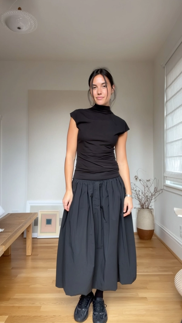 A candid, spontaneously captured iPhone photo of a woman standing still inside a minimalist Japandi-inspired apartment characterized by clean lines, warm wood flooring, and soft neutral walls. She wears a sculptural black top with a high neck and cap sleeves emphasizing minimalist elegance, paired with a stiff cotton voluminous A-line maxi skirt creating a structured silhouette, complemented by heavy black sneakers adding a subtle contrast of form and function. Soft, diffused daylight filters gently through a large window casting natural shadows and gentle highlights, illuminating her natural skin textures and subtle fabric grains. Her pose is direct and relaxed, hands naturally resting by her sides, with hair casually styled in loose strands softly framing her face with bare natural makeup, showcasing subtle skin details. The scene includes minimal décor elements such as a simple wooden bench, textured ceramic planter with dried branches, and unobtrusive framed artworks. The casual framing is slightly tilted with an intuitively spontaneous angle, reinforcing the genuine intimacy and realism seen in true iPhone candid captures.