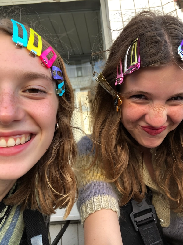 Mismatched plastic and metal clips cluster in vibrant hues along one side of each friend’s hair, catching scattered afternoon café window light as they beam with chaotic joy. The clips glint unevenly against tousled strands, while their playful expressions—one with a bright smile, the other with a mischievous pout—feel lively and candid. Soft fly-away hairs frame dewy skin, freckles peeking through natural makeup, as a knit sweater's slight fuzz presses under a chin. The shot tilts just a hint, mimicking their spontaneous lean toward the camera, hinting at a tiled café wall blurred behind. Seatbelt straps of a nearby bike messenger bag add a subtle urban touch—authentic iPhone close-up, statement hair-clip focus