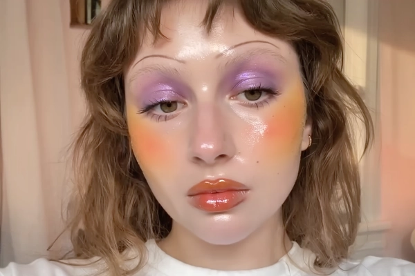 A close-up iPhone-style portrait of a young woman with natural, soft-textured hair framing her face casually. Her skin glows with a dewy, glass-like finish highlighting her cheekbones with a realistic glossy sheen and visible skin texture including pores and faint peach fuzz. Her eyelids are adorned with rich purple shimmer eyeshadow that catches natural light softly, blending seamlessly with subtle pastel undertones. She wears heavy orange-tinted blush extending across her cheeks and beneath the eyes, creating a vibrant yet natural flush. Her lips are lacquered in a high-shine translucent coral gloss that reflects ambient light authentically. Her eyebrows are natural dark brown, brushed upward lightly. The framing is tight, capturing from just above the forehead to the upper chest, with a slight tilt creating casual spontaneity. The background is softly blurred in warm, pastel peach and cream hues suggesting an indoor setting with soft window light mingled with subtle phone flash that casts natural shadows and highlights. Fine details like individual lashes, the texture of the lip gloss, skin pores, and faint flyaway hairs add tactile realism, creating the impression of a candid, heartfelt moment captured on an iPhone.