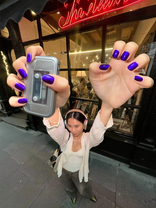A spontaneously captured iPhone-style image featuring a tiny, stylish young woman with bold purple nail art artistically reaching dramatically oversized giant hands toward the camera, holding a vintage cassette player and retro headphones. She wears a casually elegant contemporary outfit blending soft, refined textures with subtle vintage flair, alongside delicate modern jewelry and a relaxed, confident hairstyle. Behind her, a neon-lit storefront glows warmly at dusk, casting moody, atmospheric light with tasteful lens flares. Soft, natural lighting authentically highlights the realistic skin textures, detailed fabric weaves, vivid nail polish shine, and subtle jewelry finishes. The composition uses a slightly tilted, close framing creatively emphasizing the exaggerated giant hands stylishly extending forward, capturing the playful yet effortlessly chic, authentic aesthetic typical of candid iPhone photography in trendy urban nightlife scenes.