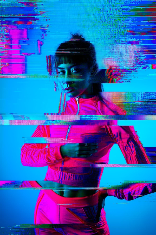 A striking female model clad in vivid bright pink sportswear stands sharply against a luminous blue wall, her figure bathed in neon saturation. Her face, partially obscured by subtle glitch artifacts, reveals eyes brimming with quiet determination, lips slightly parted, capturing a moment of introspective energy. Vertical and horizontal scan-line distortions ripple over her form, fragmenting parts of her body into pixelated shards reminiscent of corroded digital information. The background mural appears as a glitchy cascade of distorted projections, dissolving and reassembling like a corrupted low-resolution game texture; RGB color separation intensifies the surreal effect with electric blues and magentas bleeding into each other. The composition is tightly framed around the model's upper body, emphasizing the fractured blend of organic human form and mechanical disruption. Textures invoke the tactile sensation of grainy CRT scanlines and faint analog static, embedding a nostalgic digital decay. Overall, the mood is an evocative fusion of cyberpunk vibrancy and melancholic digital alienation, where human vitality clashes and harmonizes with technological breakdown, channeling themes of identity and fragmented reality in a retro-futuristic glitch aesthetic.