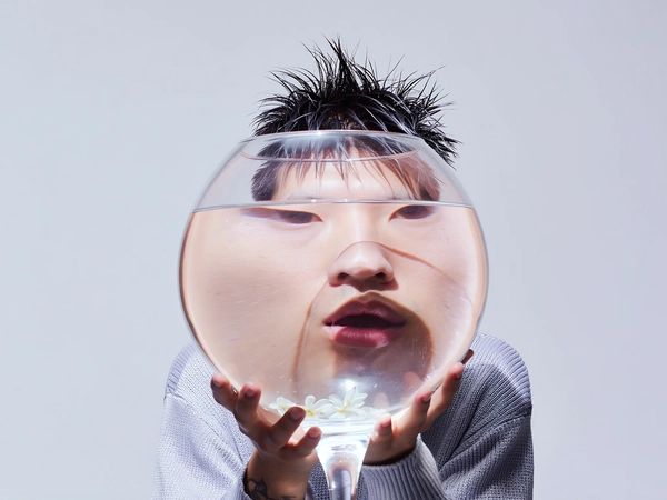 A surreal minimalist portrait of a young man indoors with spiky wet hair styled messily, embodying a textural personalization look through a light gray textured knit sweater layered casually. His face is completely obscured by a transparent glass fishbowl fully filled with clear water containing gently floating delicate flower petals, held firmly yet naturally by his own hands directly in front of his face. His slightly parted lips and vacant expression evoke a subtle post-internet alienation mood. The background is an even, soft pastel gray with no distractions. Natural soft daylight illuminates the scene, casting delicate reflections and subtle refractions through the water and glass, emphasizing authentic skin textures, natural hair strands dampened by moisture, and intricate optical distortions that elongate and duplicate facial features. The composition is casually framed with spontaneous slightly off-center alignment, preserving candid realism while highlighting surreal distortion induced by the fishbowl’s water-filled transparency, all rendered in muted neutral tones and gentle pastel grading evocative of authentic iPhone capture aesthetics.