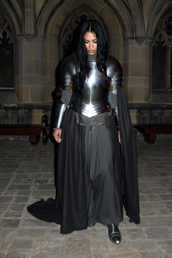 A wide, full-body, eye-level shot captured with an early 2000s-style digital camera, featuring a striking woman with long black hair and strong facial features standing indoors in a dimly lit, gothic stone interior with stone benches and high arched windows. She wears a shiny medieval chest plate paired elegantly with a dark, flowing cape over refined medieval-inspired attire. Her posture is still and reverent, with her head slightly bowed in solemn focus, eyes cast downward. Soft yet harsh direct flash typical of early 2000s digital cameras illuminates her armor's subtle reflections while leaving deep shadows that wrap around her figure, creating a moody, intimate, and raw atmosphere. The textures of her armor, natural skin, and hair strands are authentically detailed with visible digital noise and slight over-sharpening, emphasizing a gritty yet elegant medieval-core aesthetic blending medieval knightly dignity within a subdued gothic setting.