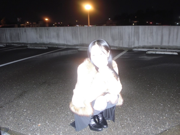A spontaneous nighttime iPhone flash photo of an East Asian woman crouching on wet pavement beneath a flickering parking lot lamp. She wears a fur-trimmed coat, long skirt, and heeled boots, but the intense direct flash overexposes her completely, bleaching out facial features and clothing details, leaving only ghostly, luminous outlines. The cold concrete environment is dimly visible in the background, framed with the imperfect, casual angles typical of candid iPhone snapshots, creating a surreal and ethereal atmosphere.