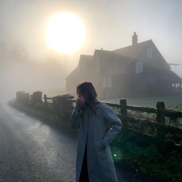 A young woman clad in a sleek, minimalist trench coat stands still on a slick, rain-dark asphalt path winding past a rustic cottage whose outlines blur softly into thick, lingering fog. Her loose hair clings damply to her neck as she tilts her chin slightly forward, breath visible in the cold, and one hand slides into her coat pocket while the other brushes a stray strand from her face. The dense mist swallows color into pale greys and muted greens of moss clinging to gateposts, while a faint, diffused dawn light softly pools through the haze, casting gentle luminance with a subtle lens flare near the blurred sun. The iPhone's grain texture whispers through shadowed corners; droplets bead quietly on wilting leaves, and the faint tap of distant dripping condensation layers the stillness. The shot, caught from chest height with a hand-held, slightly tilted frame, leaves generous negative space above and around her, dissolving the world into quiet solitude—mistbound reverie, shot on iPhone