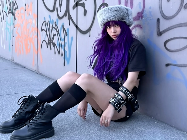 A candid mid-2010s style photograph of a young woman with vivid deep purple tousled waves, sitting casually on the floor leaning against a graffiti-covered urban wall. She wears chunky black combat boots, black knee-high socks, and a cozy pale gray faux fur hat, complemented by studded leather cuffs adorning her arms. Her expression is a genuine unimpressed death glare toward the camera. The scene is illuminated by soft natural outdoor daylight with intentionally muted saturation and softened contrast levels, while shadows carry a cool-toned lavender-gray hue. Visible textures include the rough fabric of her socks, the rugged leather boots, detailed studded accessories, and realistically defined loose strands of her vibrant hair. The photograph features a slightly tilted, off-center framing with casual cropping, evoking spontaneous authenticity and relaxed intimacy consistent with mid-2010s candid youth photography aesthetics.