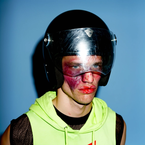 He’s framed in a medium close-up against a matte baby blue studio backdrop, the shot uncomfortably hand-held with slight asymmetry. His face is partially obscured by a scratched, glossy black motorcycle helmet with a slightly fogged visor that reflects the harsh studio lighting. His cheek is bruised in deep purples and bruised reds, lips swollen and glistening like a prizefighter post-bout, yet his deadpan stare pierces through the visor with icy electric blue eyes. He wears a black mesh shirt under an unzipped neon green sleeveless hoodie, the collar peeking out just enough to clash with the bruised softness of his features. The whole moment holds a violent yet glamorously unbothered tone, like a Y2K skater who’s just stepped off a backstage ramp ready for the flash. The lighting casts soft shadows and mild flash glare on the visor’s scratches, emphasizing texture and raw grit.
—casual candid early-2000s Y2K snapshot, grainy softness