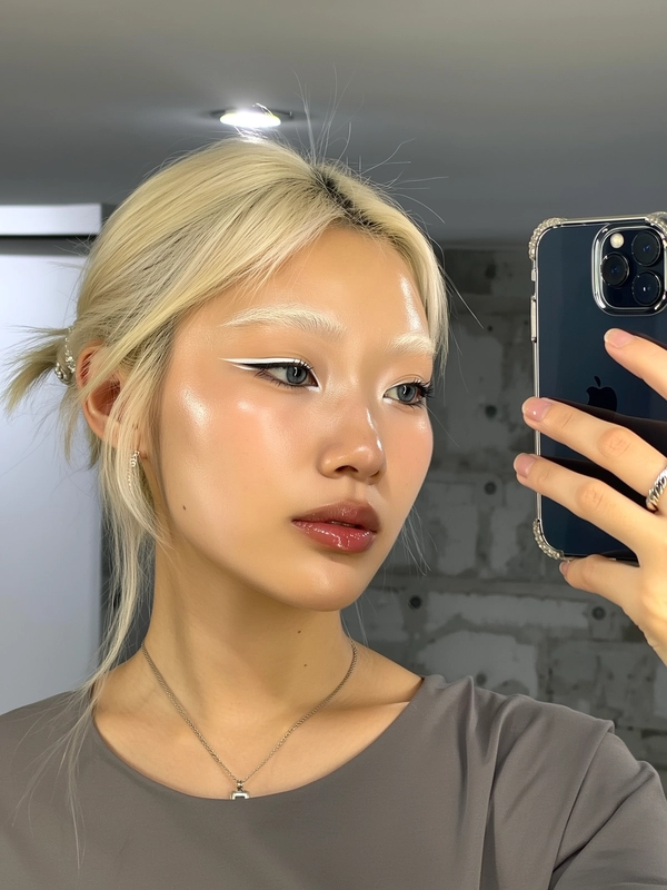 A natural close-up iPhone selfie of a young woman with naturally bleached eyebrows that gently blend into her skin, creating a subtle pale blonde effect. She features striking silver-gray eyes subtly enhanced by dramatic but refined white eyeliner, applied with a clean and natural finish. Her skin appears luminous and glassy with realistic texture and soft natural gloss. The expression is calm and engaging, looking directly into the camera with a neutral yet captivating gaze. She wears a simple muted gray top with minimalistic silver jewelry. The setting is a minimalist, brutalist-inspired indoor space subtly lit by a single soft overhead light creating gentle shadows, emphasizing the authentic handheld selfie aesthetic with neutral, textured background elements.