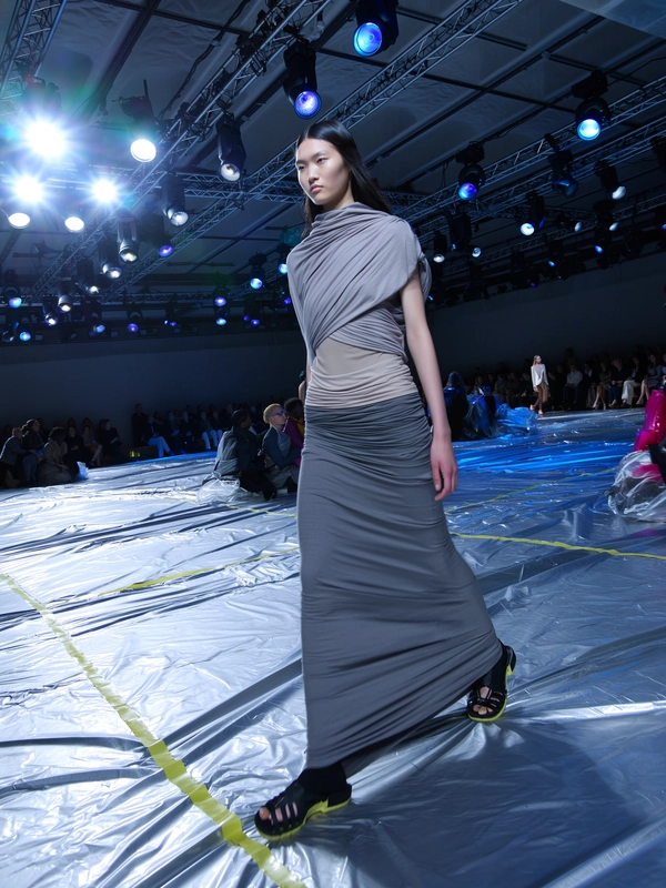 A model draped head-to-toe in muted greys and soft taupes strides confidently through a runway lined with crumpled metallic sheets that shimmer like lunar rock under a cool, soft blue backlight. The silhouette is loose yet sculpted, featuring layered twisted fabrics and matte textures that absorb the eerie glow. Her expression is calm, almost otherworldly, blending into the surreal, moonlit set where scattered audience faces lean forward on minimalist benches. Above, industrial trusswork frames rows of blue-tinged spotlights and tungsten bulbs, casting a mix of sharp halos and gentle shadows that the iPhone sensor captures with mild digital grain and a slight bloom around the brightest light pools. The runway’s metallic floor bears taped sight-lines and subtle scuffs, while the model’s skin shines faintly beneath the cool light, and a gentle motion blur kisses the sweeping hemline mid-stride. Shot from a low, angling aisle perspective with a blurred edge and soft hand-held shake, the scene pulses with dreamy editorial tension—contemporary runway snapshot, shot on iPhone