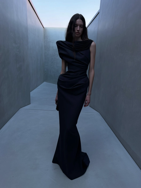 A softly dim-lit iPhone snapshot capturing a model dressed in a floor-length black gown by Rick Owens, featuring structured sculptural draping and an asymmetrical neckline with gathered fabric layered over one shoulder. She stands slightly off-center within a narrow, angular Brutalist corridor characterized by cold, muted grey concrete walls devoid of direct sunlight. The cool dusk-inspired ambient lighting gently diffuses over the matte textures of the fabric and the subtly worn concrete, highlighting delicate folds and architectural geometric forms. The composition is elevated and tilted downward subtly, framing the model with generous negative space and muted shadow gradients, evoking an intimate, quietly moody evening moment reminiscent of a spontaneous, minimalist fashion capture on iPhone.