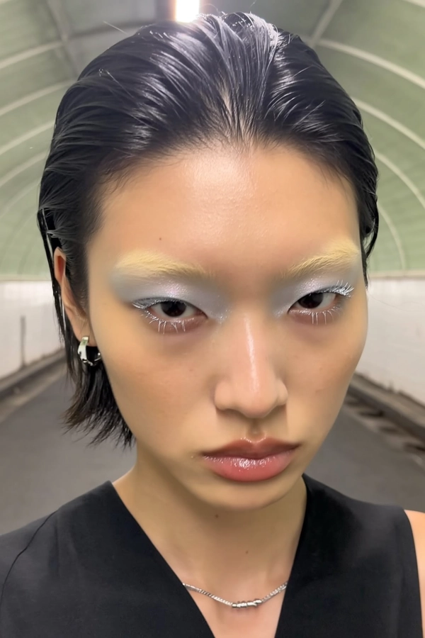 A natural close-up iPhone selfie of a young model with naturally bleached eyebrows that blend softly into her skin, effectively appearing almost absent, paired with subtle silver eyelashes that add a delicate shimmer to her eyes. Her slick, wet-look hair is styled back neatly, framing a vacant but calm expression as she engages directly with the camera. Minimalist makeup highlights muted, slightly glossy nude lips and an icy silver eyeshadow that complements the lashes. She wears a simple black top with subtle texture, accessorized only by minimalistic silver jewelry. The background features a softly lit, neutral-toned underground tunnel with subtle textures, maintaining an authentic and effortlessly cool urban vibe typical of candid iPhone selfies.