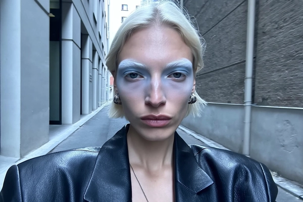 A close-up, authentic iPhone selfie featuring a sharp-cheekboned model with naturally bleached eyebrows that subtly blend into her pale skin, creating a soft, pale blonde effect. Her makeup focuses on icy blue eye shadow that delicately enhances her eyes, paired with muted, natural-toned lips. She wears a simple black leather coat reflecting contemporary, effortless style, with minimalistic silver jewelry. Her expression is neutral, directly engaging the camera with a calm gaze. The background shows a narrow urban alley framed by brutalist towers with textured, neutral concrete surfaces. The lighting is slightly harder with a cool blue tone, realistically illuminating her facial contours and makeup while preserving natural skin texture and slight imperfections, capturing the candid, stylish, and casual essence of an iPhone selfie.