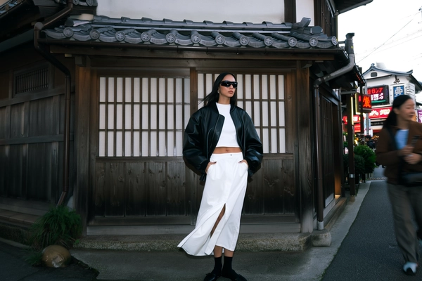 Leaning statuesque against a weathered wooden facade beneath a traditional tiled roof, a confident woman draped in a sleek black leather bomber jacket slips her hands casually into pockets above a white cropped top. Her high-waisted white maxi skirt with a bold front slit flutters slightly in a gentle Tokyo breeze, revealing just a hint of legs; minimalist black shoes ground her cool poise. Futuristic wrap-around sunglasses shield sharp eyes as dusky daylight dims into softly glowing streetlights. The textured grain of aged wood and sliding doors contrasts sharply with modern leather sheen and crisp fabric folds, framed off-center with gentle lens flare bouncing from nearby neon signs. A faint blur of slow-moving passerby merges with the serene vintage backdrop, balancing heritage and streetwear edge in a raw, handheld moment near Kyoto-style alleys touched by Tokyo style. —Tokyo street snap, shot on iPhone
