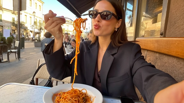 Under the warm, golden-hour glow of a late afternoon in Milan, a brunette woman sits poised at a stylish sidewalk café. Her hair is smooth and meticulously parted, framing her face with quiet sophistication. She wears a tailored, navy blazer that catches the soft light, paired with classic dark sunglasses that rest lightly on her nose, giving off an effortless high-fashion aura. Her lips, subtly glossy, catch the eye with a faint blush of rich red sauce, a delicate mark of her enjoyment.

With precise grace, she lifts a glossy forkful of spaghetti dripping with thick, aromatic marinara, the tendrils curling artfully as she draws it toward her mouth. Her eyes, visible just above the rims of her sunglasses, soften in calm focus; lips part slightly around the pasta in a moment caught between thoughtful and playful. The sauce’s texture glistens faintly, the rich sheen contrasting with the crisp fabric of her blazer.

Around her, the quaint sidewalk buzzes gently—the muted chatter of fellow diners and the distant hum of life weaving through sunlit cobblestones. The café table's textured wood grain is faintly visible at a casual arm’s-length selfie angle, tilted just so, capturing the scene’s intimacy and understated elegance.

The soft amber hues and natural backlight carve subtle shadows that accentuate the integrity of her poised form, immaculately styled yet relaxed. This serene, lightly candid image balances the casual pleasure of eating with a refined, editorial presence—perfectly resembling a candid iPhone photo.