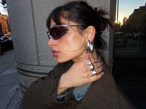 In a raw city corner washed with dull afternoon light, a woman in a dark beige coat presses her hand behind her neck, revealing chunky silver rings that flow like liquid metal, their molten curves catching the faint lens flare from nearby glass. Her profile tucks just out of focus, a few stray hairs slipping from a messy bun, while large sculptural earrings glint softly. The iPhone's center-weighted focus blurs the gritty backdrop, denim collar fuzz just noticeable as it frames skin pores and a tiny lens smudge. The frame tilts, emphasizing her aloof gaze and the half-hidden play of shadows—the urban gloss reflected dimly in wrap-around sport shades. —close-up captured on Iphone, hand-face jewelry focus