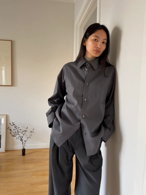 A spontaneous, casually candid iPhone image of a woman leaning lightly against a warm-toned wall near a minimalist doorway in a Japandi-inspired home interior. She wears a sharply structured charcoal button-up shirt with a high collar layered minimally over a subtle textured neutral undershirt, paired with full-length straight-leg trousers in matching dark charcoal fabric. Her hands rest naturally in her pockets, and her expression is composed and almost blank, reflecting quiet introspection. Soft natural indoor daylight gently illuminates the scene, casting diffused shadows enhancing the textures of the sculptural fabric and smooth skin with subtle imperfections. The background includes muted wooden flooring, simple framed minimalist artwork, and a single dried branch arrangement, lending a domestic yet editorial Japandi ambience. The framing is casual with a slight tilt capturing the effortless relaxed pose and intimate atmosphere typical of authentic iPhone candid photography.