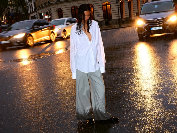 She stands mid-street beneath a damp evening sky, oversized white shirt clinging with fine rain beads to smooth sleeves stamped with faint text, the wide light-gray trousers darkened by soaking puddles at the hems. Her wet hair clings softly around a calm, downward-tilted face, relaxed posture leaning gently to one side. Behind her, a grand beige stone façade with ornate windows frames the quiet drama, while warm golden car headlights scatter across slick asphalt that mirrors each light in rippled pools. The blend of cinematic golden glows and cool concrete dusk balances softness and edge, with drifting raindrops caught speckling the lens. The handheld eye-level perspective tilts subtly, the street's reflective sheen rippling under footfalls beyond the frame, blending calm and motion in a perfect rainy blur—rainy city snap, shot on iPhone