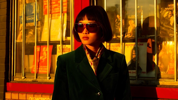 In the dusky glow of a fading afternoon, an East Asian woman stands statuesque in front of a weathered record store, the peeling posters behind her whispering stories of bygone concerts and local legends. Her sharp cheekbones catch the amber sodium-vapour streetlight at an angle, casting soft shadows that sharpen her dramatic gaze beneath oversized tinted sunglasses, their smoky lenses refracting diffused multicoloured neon glints. Draped in a deep green corduroy blazer, the fabric’s distinctive ridged texture is palpable under the warm shadows, while a silk scarf with subtle paisley patterns wraps elegantly around her neck, the sheen catching flickers of muted sunlight. The scene is bathed in a golden-hour Kodachrome hue, saturating every fold and wrinkle in her attire, as authentic 35 mm grain and a hint of gate-weave animate the tactile quality of her skin pores and the crumpled paper of worn posters. Captured at eye level through a 50 mm lens, the composition frames her with calm authority—shoulders squared, weight evenly balanced—immersed in an unmistakably vintage urban tableau. The worn brick façade, metal-framed storefront windows, and dim glow from inside the store complete the ambience, evoking a candid moment of poised strength in the late-70s cityscape. —late-70s / early-80s cinematic photograph, authentic film grain.