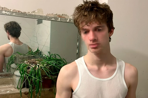 Casual candid image of a pale young man standing indoors in front of a cracked mirror and a dying houseplant, wearing a plain white sleeveless tank top. His face is deliberately pixelated to preserve anonymity and add visual intrigue. The background features a peeling wall with authentic texture details. Soft, natural indoor ambient lighting typical of iPhone photography gently illuminates the scene, capturing realistic fabric textures of the tank top, natural hair strands, and subtle imperfections of the environment. The framing is informal and slightly tilted, reflecting spontaneous, unscripted capture.