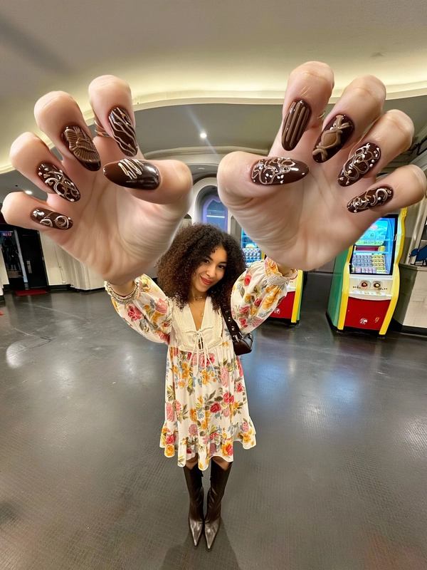 A spontaneously chic iPhone-style image featuring a tiny, fashionably dressed curly-haired woman wearing a vibrant floral dress and sleek high boots, casually extending dramatically oversized, elongated giant hands toward the camera. Her nails showcase intricate hand-painted chocolate-themed designs with realistic glossy textures. The background reveals a softly lit retro arcade environment with gently diffused ambient indoor light creating natural highlights and nuanced shadows. The composition captures a slightly tilted, wide-angle framing that enhances the exaggerated proportion of her giant hands, while authentic skin imperfections, detailed fabric textures of her floral dress, refined leather of the boots, and subtle reflections contribute to sophisticated textural realism. Her relaxed yet confident expression and effortless posture embody a playful and elegant casual style emblematic of spontaneous, fashionable iPhone photography.