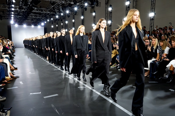 Twenty statuesque models sweep down the runway in a tight line, each cloaked in sleek, all-black ensembles ranging from sharp tailoring to flowing asymmetrical silhouettes. Their expressions, fierce and unified, cut through the crescendoing soundtrack as sequins flicker and matte fabrics absorb the spotlight, creating a rich monochrome tapestry. Flashes erupt from hundreds of cameras held aloft like strobe-lit constellations, momentarily illuminating rows of enthralled spectators seated along the industrial-style venue, complete with exposed steel trusses and high-tech LED rigs. The catwalk’s matte black floor bears scuff marks and duct tape lines leading the way, catching faint reflections of each stride. The iPhone’s sensor softens bursts of light into radiant halos while catching subtle motion blur on trailing hems and confident steps, with a slight handheld wobble that lends visceral immediacy. Shot from just off-center in the first row, the framing slices asymmetrically through the crowd, their whispered excitement tangible in the capture—contemporary runway snapshot, shot on iPhone