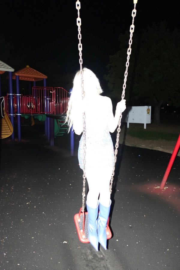 A spontaneous iPhone snapshot at night on a playground, capturing an Afro-Latin woman standing on a swing mid-motion. The intense direct flash overexposes her completely, bleaching out all facial features and details of her glittery mini-dress and high boots, leaving only a glowing, ghostly silhouette. Her playful and defiant pose is frozen in imperfect, candid framing, surrounded by the dark outlines of playground equipment, creating a surreal and abstract visual effect.