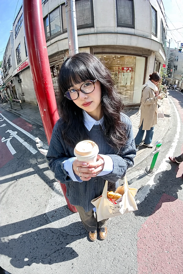 A bright midday natural light wide-angle iPhone-style photo with slight fisheye distortion, capturing an elevated view of a stylish young woman standing on a narrow urban Tokyo sidewalk. Her long wavy black hair frames a face wearing unique tortoiseshell-rounded eyeglasses with thick frames. She wears a dark grey textured cable-knit sweater layered creatively over a pale blue button-down shirt with an artfully visible collar, merging casual and preppy styling with original details like subtle knit patterns and slightly relaxed sleeve cuffs. Both hands hold an imaginatively designed light beige takeaway coffee cup with a matte plastic lid, raised gently toward the camera. She tilts her head naturally to one side and looks directly up into the lens with a neutral, subtly observant expression, conveying authentic engagement without smiling. Her footwear are original metallic bronze chunky sneakers featuring soft gleaming leather textures distinct from referenced styles. Nearby at her feet rests a cream-hued canvas tote bag, decorated with a novel printed abstract design, filled with bakery-style items casting soft realistic shadows on worn asphalt. The street scene features quintessentially Japanese urban elements: a small commercial building with reflective glass windows, authentic kanji signage, and a vivid red vertical pole partially cutting through the left frame edge. The background includes two slightly blurred pedestrians dressed in creatively reimagined neutral-toned coats—one in a soft beige layered with a white scarf, the other in taupe with stylish sunglasses—emphasizing candid street life. The pavement is weathered asphalt with white lane markings and green-and-white safety barriers; intricate shadows from a metal railing cast dynamic diagonal lines adding depth. Natural bright sunlight creates realistic highlights and shadows, accentuating the varied textures of clothing fibers, skin details, hair strands, and urban surfaces. Compositionally, the image uses a thoughtfully elevated tilted fisheye angle typical of casual iPhone photography to exaggerate perspective dynamically, emphasizing playful yet authentic distortion that enlarges the upper body and foreshortens feet, centrally framing the engaged subject. This spontaneous candid wide-angle shot balances a stylish, curated look with natural urban spontaneity, embodying a distinctly modern, intimate street portrait.