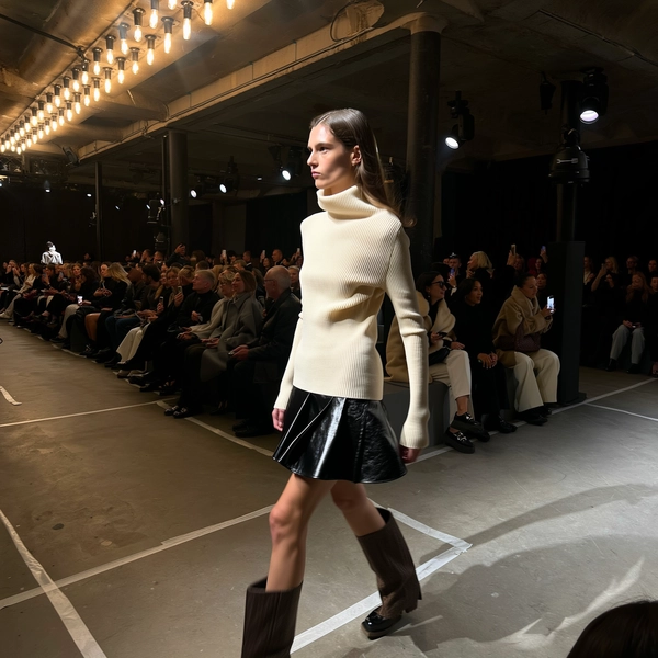 A poised model commands the runway wearing a soft cream knit top that hugs her torso with delicate ribbed texture, paired with a glossy black leather mini skirt that sways with each confident step. Her regal posture radiates statuesque elegance as she strides past a muted gray-clad audience seated on minimalist benches, their faces shadowed yet engaged. The industrial venue’s concrete floor is marked with scuffed taped sight-lines, while overhead rows of tungsten can lights cast warm pools alongside colder LED spotlights, creating a subtle interplay of shadows around her silhouette. Slight motion blur trails the leather hem and the gentle swing of her arms, while the iPhone lens picks up the soft sheen on her skin and fine knit weave amidst faint sensor noise in the dimly lit corners. Shot from an oblique front-row angle, the frame tilts just enough to convey handheld immediacy, capturing whispered guest reactions and raised phones at the edge of the frame—contemporary runway snapshot, shot on iPhone