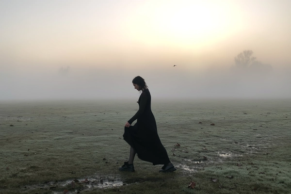 A solitary woman draped in a flowing black dress moves slowly across a dew-soaked open field, the morning mist blurring the distant horizon into soft, pearly greys. Her dress sways with each careful step, hints of damp wool fibers and loosened hair strands brushing against her neck caught in the cool, veiled air. The low fog softens the pale dawn light, casting muted shadows and a faint halo around the rising sun. From a waist-high, handheld vantage, the image trembles slightly with the subtle motion of her movement, wide negative space amplifies her lonely passage through the slick grass and muted earth tones. A faint rustle of wet leaves and a distant crow’s call underpin the stillness, the whole moment wrapping in quiet introspection and mistbound reverie, shot on iPhone.