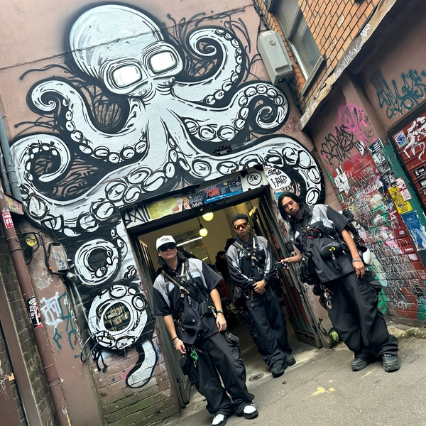 A candid street photo of three friends casually posing in coordinated black and gray techwear outfits with layered street style, harnesses, and utility accessories, standing relaxed in a narrow urban alley. Behind them, a large, detailed graffiti mural of an octopus dominates the doorway, its eyes replaced by vintage TV heads; the octopus's tentacles twist and dissolve into glitch-like brushstrokes extending along the brick walls. The alley’s surfaces are heavily textured with peeling paint, faded posters, and layered graffiti tags. Natural daylight filters softly into the confined space, casting subtle shadows and highlighting the rough urban textures. The composition uses a slightly tilted, eye-level framing, capturing an authentic spontaneous moment typical of candid iPhone photography with genuine street ambiance, rich graffiti detail, and realistic tactile surfaces.