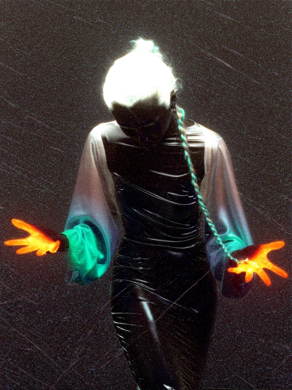 A luminous figure crouches low, one iridescent braid cascading and bleeding into the obsidian void that swallows her form; her hands splay wide, fingertips vaporising in radiant orange and electric teal pulses that streak like after-image trails across taut, melted latex attire clinging to sinew and bone. Her skin liquefies mid-motion, dissolving into streaming pastel rivulets that warp and ripple beneath a plasma-white rim light flipping hues through an infrared swap, making shadow and light twist in spectral rapture. Coarse 800 ISO grain clusters embed within the folds of her ghostly silk sleeves, dust specks fading into faint gate-weave jitter that pulses softly as radial blur trails smear off the edges of her stance. The photo quality suggests a slightly grainy or low-resolution digital look, capturing a candid moment. Shot through a Canon EOS R5 with an RF 85 mm ƒ/1.2, layered with scanned tungsten-tinted film grain, sprocket burn remnants trace the frame edges—surreal inverted-color fashion editorial, heavy film grain texture