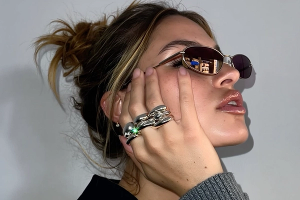 A woman with her hair loosely slipping from a messy bun tilts her head just so, a faint frosted gloss tracing her lips as afternoon light softens the clean wall behind her. Her hand presses gently against her cheek, rings stacked in bold, molten-silver shapes that seem to flow and catch tiny lens flares along their curves. Dark micro-frame shades slant down her nose, reflecting a muted cityscape just out of view. The grain of her rib-knit cuff peeks in, blending with the soft texture of skin pores and a faint lens smudge, while a slight Dutch tilt hints at a candid moment caught off-guard— close-up captured on Iphone, hand-face jewelry focus