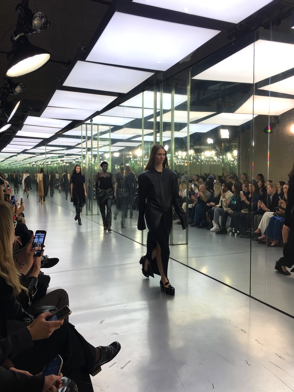 A wide-angle view down the full length of a glass-walled, mirrored runway floods with piercing LED panels that cast sharp reflections and gleams on the polished floor. Models stride with cool, emotionless visages, their avant-garde silhouettes rendered in sleek leathers and translucent fabrics, their slow-motion steps sending subtle ripples through chiffon sleeves. Audience benches flank the catwalk, blurred hands holding phones aloft while a soft murmur permeates the air. Industrial fluorescents blend with tungsten rigs overhead, creating crisp highlights and softer spill illuminating the mirrored walls. The iPhone sensor picks up slight digital noise in the shadowed corners and a delicate bloom around the brightest fixtures. Faint scuff marks and taped sight-lines mar the runway’s glossy surface, anchoring the moment’s organic grit. The shot is taken from a raised rear seat, a slight diagonal tilt capturing the full geometry of the space and the linear power of the procession—a natural street-side snapshot pulsing with editorial energy—contemporary runway snapshot, shot on iPhone