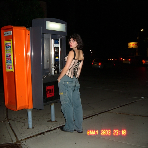 The flash pops hard against the dim streetlight, casting the woman in her vintage corset top and baggy cargo pants into a stark silhouette beside the beep of the old payphone booth. The tungsten glow bleeds orange along the cracked pavement, while faint blocky JPEG artifacts crunch the dark edges of her form. Her eyes glance just above the lens, caught mid-thought as the timestamp flickers in the corner—"04 MAR 2003  23:18"—adding to the lost-movie feel. The image has a slightly blurry and low-resolution quality, resembling a video still or casual recording, with grainy speckles dotting shadow patches and soft fading around the crooked tilt of the frame. Battery icon blinks weakly near the date, as if the night itself is winding down—early-2000s digicam snapshot, grainy low-res quality