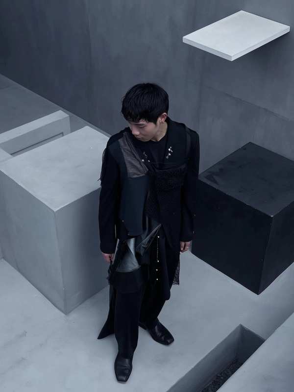 A quietly moody iPhone snapshot capturing a man dressed in an avant-garde outfit inspired by Junya Watanabe Comme des Garçons menswear, featuring layered black fabrics with mixed textures including mesh and shiny materials, creating a voluminous, architectural silhouette. He stands slightly off-center within a softly dim-lit Brutalist concrete setting characterized by angular geometric forms reminiscent of black cubes and a subtle floating white rectangular shape. The lighting is cool, dusk-inspired with muted grey and charcoal tones, softly diffused to reveal matte fabric textures and quiet imperfections in the concrete. The composition employs a gently elevated, slightly downward angled view, framing the model with generous negative space and subdued foreground elements, conveying calm introspection and effortless intimacy.