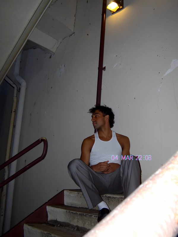 The dull tungsten glow from a streetlamp catches the sharp crease of his tailored trousers as a sudden flash from the camera stutters across the ribbed tank top. He sits alone on a worn building staircase, gazing quietly off to the side, the edges around him soft and a little smeared with CCD noise speckles. The built-in flash casts harsh shadows that bleed slightly, merging with faint JPEG blocks on the wall’s chipped paint. The angle’s slightly tilted, like a hurried zoom caught mid-shake, while a pale blue timestamp reading “04 MAR 2003  23:18” blinks faintly in the corner. The photo quality is low-resolution and somewhat grainy.—early-2000s digicam snapshot, grainy low-res quality