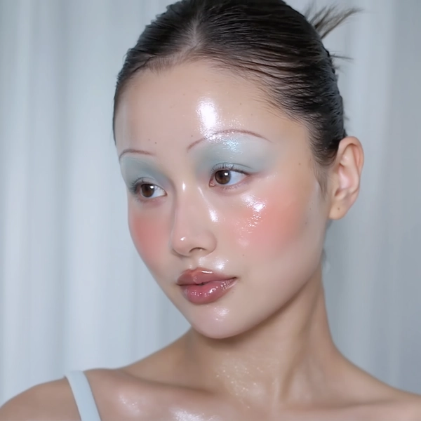 A close-up iPhone-style portrait of a young woman with tightly pulled-back hair revealing her natural hair texture and subtle flyaways, styled simply without overly slick elements. Her skin features a luminous, dewy finish with visible pores and soft natural sheen across her forehead, nose, and cheeks. Makeup channels a gentle babydoll aesthetic with neon-inspired pink blush richly diffused across her cheeks and softly extending beneath her eyes, paired with shimmering pastel eyeshadow in soft icy blue creating a subtle glow on her lids. Her lips wear a glossy pale pink tint, reflecting light naturally with moist texture. Her eyebrows are natural dark brown, brushed upwards lightly without bleaching. The photo is framed tightly from just above her head to below her collarbones, captured at a slightly off-center, casual angle to replicate a candid iPhone portrait. The background is softly blurred in cool pastel tones with hints of a softly textured curtain or wall, giving a muted futuristic atmosphere but remaining minimal and non-distracting. Lighting is natural window daylight mixed with faint soft phone flash, producing believable highlights on her glass-like skin and delicate shadows along her neck and collarbones. Fine textural details like individual hair strands, skin pores, gloss reflections, and fabric weave are clearly visible, supporting a genuine, intimate, spontaneous feel.