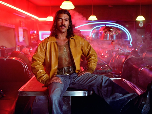 In a smoky late-70s diner bathed in glowing red neon, a Latino man lounges nonchalantly across a vinyl booth, his long wavy hair flowing casually over the gleam of a classic handlebar mustache. His mustard-hued leather jacket, slightly worn and supple, slips open to reveal a daring mesh shirt that clings softly, catching the lingering neon glints. His flared jeans fan out, edges kissed by the soft sheen of the diner’s chrome-plated tables and curved seats that frame him perfectly.

The lighting is a seductive cocktail of warm sodium-vapour lanterns mixing with the electric ruby pulse from neon strips, lending a moody, saturated glow to his sharp yet relaxed gaze. His skin displays a subtle texture of pores and light beard shadow, accentuated by the tactile grain of 35 mm film, which drapes the image in a gently scratched matte finish typical of Kodachrome stock from the period. This lends an organic, tactile quality to his confident stance.

Shot from eye-level with a 50 mm lens, the composition centers tightly on the subject's upper body, capturing the vivid textures of the leather and mesh alongside blurred chrome reflections. The diner’s sprawling jukebox silhouette occupies the background, deepening the frame with its retro-futuristic curves. Sharp shadows and warm highlights create an interplay that recalls the visual vocabulary of cinematographers like Gordon Willis.

The overall effect is unmistakably vintage—a narrative frozen in sumptuous reds and glints of silver, a portrait of relaxed cool amid the electric heartbeat of an iconic American diner. An effortless blend of intimacy and style, caught on authentic film with that rich analog grain texture.

—late-70s / early-80s cinematic photograph, authentic film grain.