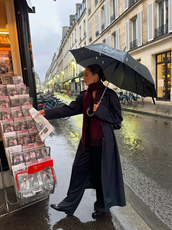 Under the soft drizzle by a corner newsstand on a quiet Parisian street, she stands poised in a dark oversized trench coat with shoulders lightly raised, black straight-leg trousers flowing with polished loafers just skimming wet pavement. A burgundy scarf drapes loosely around her neck, tucked slightly inside her coat, gold earrings flashing as her slick low bun catches scattered neon shards. Holding a slick black umbrella angled back, droplets bead its surface while her outstretched hand reaches for a folded newspaper amid fluttering wet magazines stirred by the misty breeze. Textured stone walls and shuttered windows rise behind rows of parked bicycles, their forms softened in reflected neon hues shimmering across the damp sidewalk. The overcast sky mutes daylight, sodium streetlamps casting halos that ripple across puddles. The handheld iPhone frame tilts gently mid-distance, catching an intimate, cinematic profile in candid pause — rain tracing fabric textures, umbrella ribs, and the subtle sheen on her face. —rainy city snap, shot on iPhone