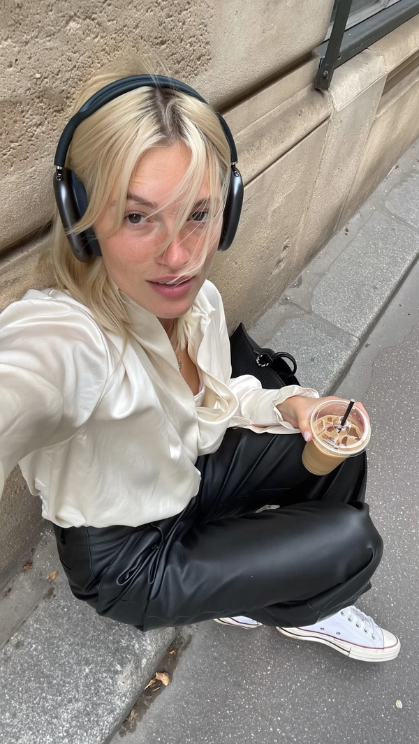 A spontaneously captured iPhone-styled candid photo of a young woman with platinum hair casually lounging against a textured, slightly weathered Parisian stone wall on a city sidewalk. She wears understated effortless cool conveyed through an ivory silk blouse from COS with soft draping and subtle fabric wrinkles paired with sleek black leather pants by Our Legacy, complemented by classic white Converse sneakers gently scuffed from wear. Over her hair sit minimal black headphones, resting lightly and adding a contemporary casual touch. In one hand, she holds a translucent iced coffee glass showing realistic condensation and subtle reflections. Her hair is styled in a naturally tousled manner, some strands catching soft natural daylight that gently illuminates her neutral makeup and natural skin texture, including faint freckles and fine pores. Surrounding her are authentic urban textures of worn concrete pavement and the rough stone wall with patches of subtle moss. The composition features a slightly tilted overhead angle, off-center framing, and an informal crop that evoke the intuitive spontaneity and genuine intimacy emblematic of everyday Parisian iPhone street photography. Soft shadows and delicate highlights reveal the delicate fabric fibers, realistic leather grain, and glass transparency, completing the true-to-life candid aesthetic.