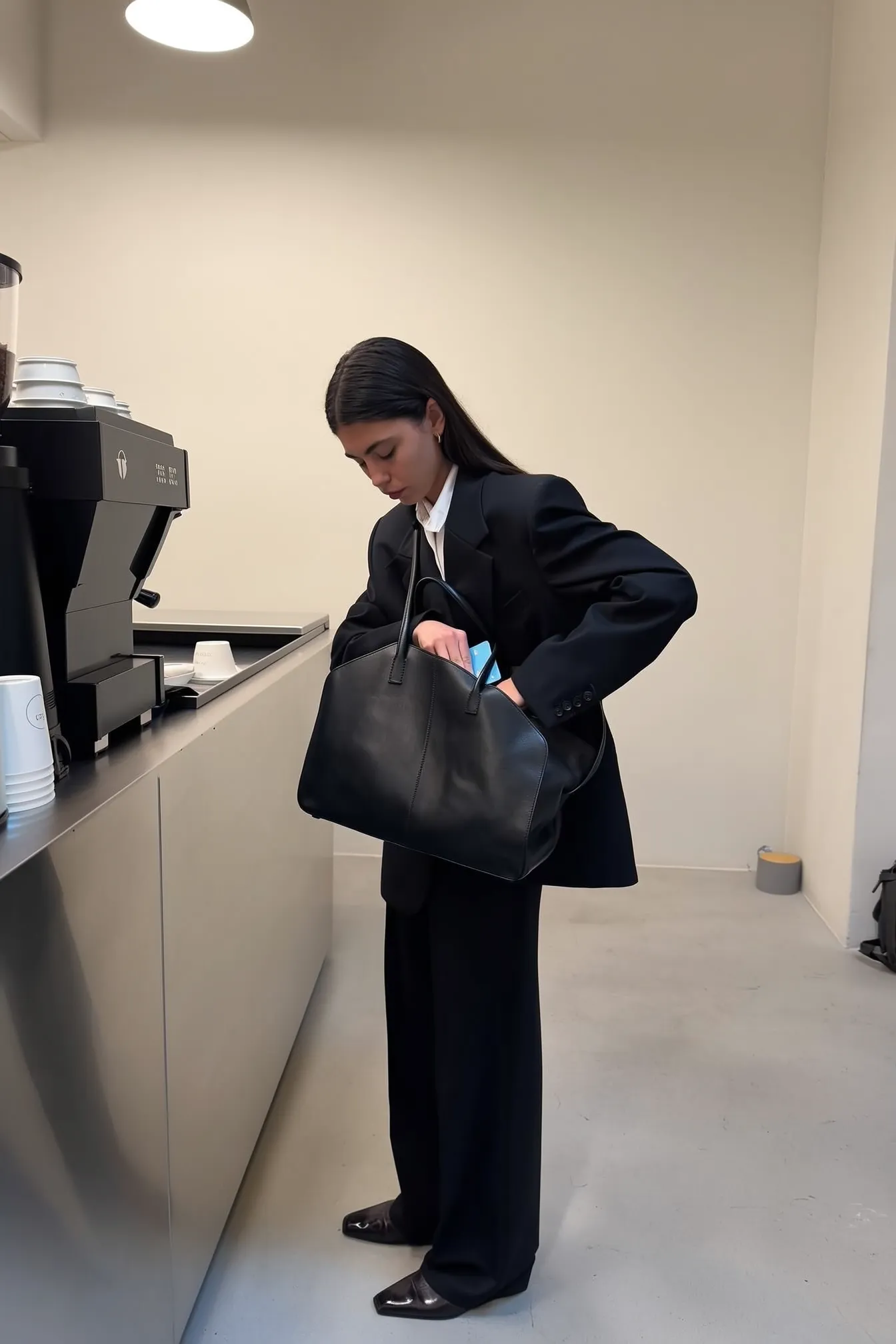 18. A girl in a business suit stands at a café counter, one hand inside her large black tote bag, searching for her card. Minimalist setting, neutral tones, and overhead lighting. iPhone perspective.