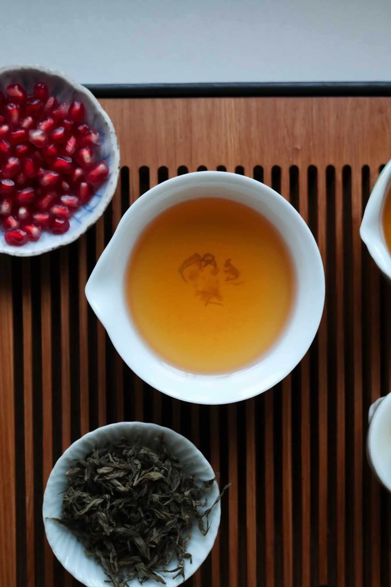 A bird’s-eye view of a wooden slatted tea tray. Centered: a shallow white porcelain teacup with golden Darjeeling tea, and a gaiwan beside it filled with spent twisted leaves. In the corner, several ruby red pomegranate seeds in a tiny ceramic bowl. Calm morning setting, subtle reflections in the tea, quiet contemplative energy.