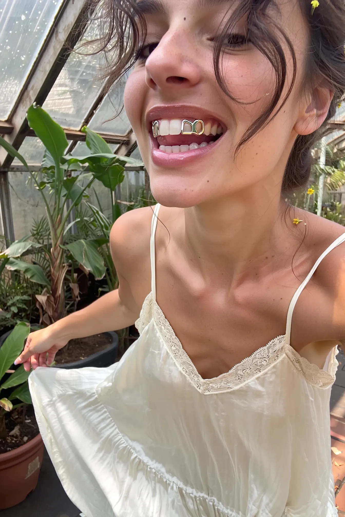 A close-up upper-body shot of a young woman caught mid-movement, on her teeth shining minimalistic, reflective metallic grills only on the upper right canine and lateral incisor, or alternatively on the lower left two premolars, with an open-frame design such as letters, stars, or abstract shapes. The rest of her teeth remain natural and white. The grills are lightweight, cleanly fitted, and realistic, either silver or gold. She wears a flowing ivory slip dress with fine lace along the neckline, the fabric softly textured and gently rippling with her spinning motion. Tiny flowers interwoven in her loosely tousled hair add a delicate touch, some strands escaping to frame her face naturally. Warm daylight streams through the overgrown tropical plants in the humid greenhouse, dust particles visible in the air, casting soft, diffused light that accentuates natural skin textures and fabric grain. The image is framed with a slight tilt and a casual, spontaneous feel, echoing the intimate authenticity and natural imperfection of a candid iPhone snapshot. The lighting, skin texture, and gloss of the lips remain exactly the same as before.